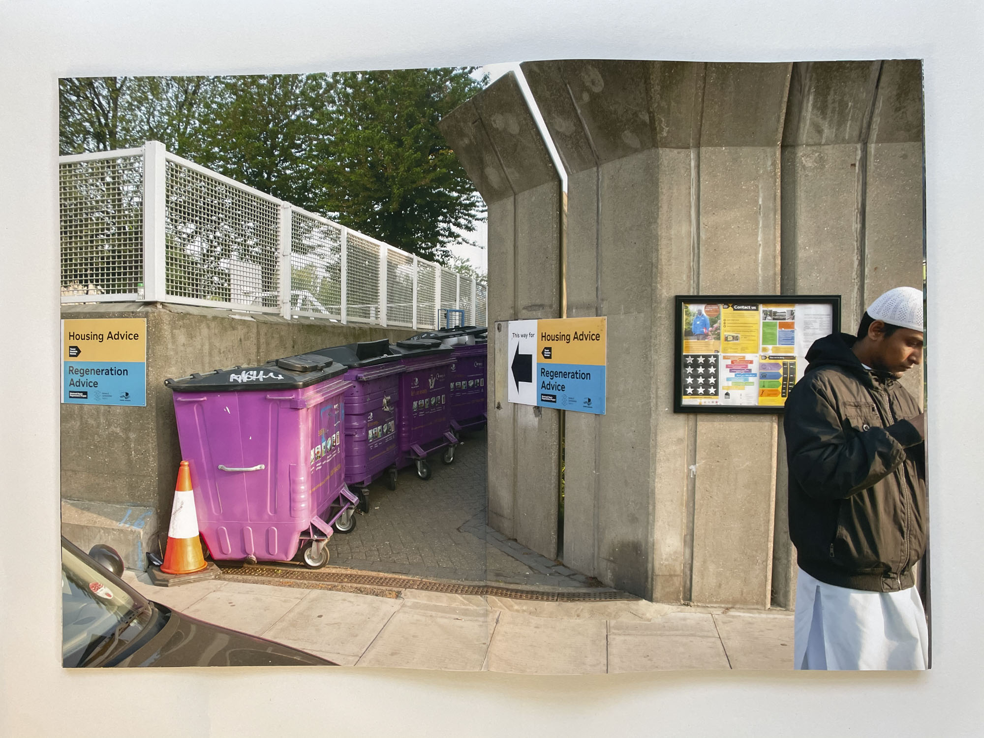 the entrance to an estate is clogged with purple refuse bins, to the right a man leaves wearing a kufi cap