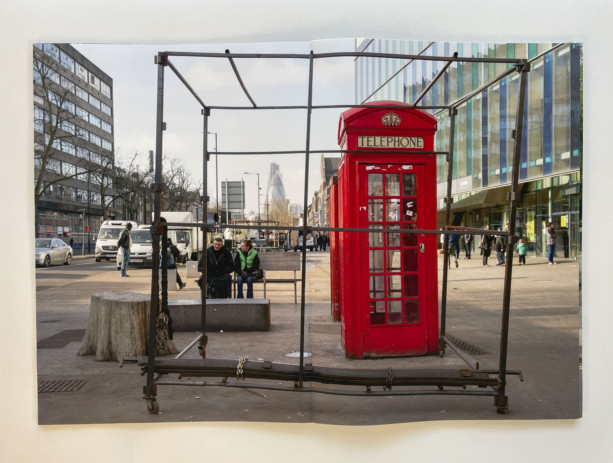 a view down Whitechapel High Street seen through the frame of a market stall, in the foreground is a red telephone box, in the middle distance an older couple sit on a bench, in the distance the Gherkin is visible