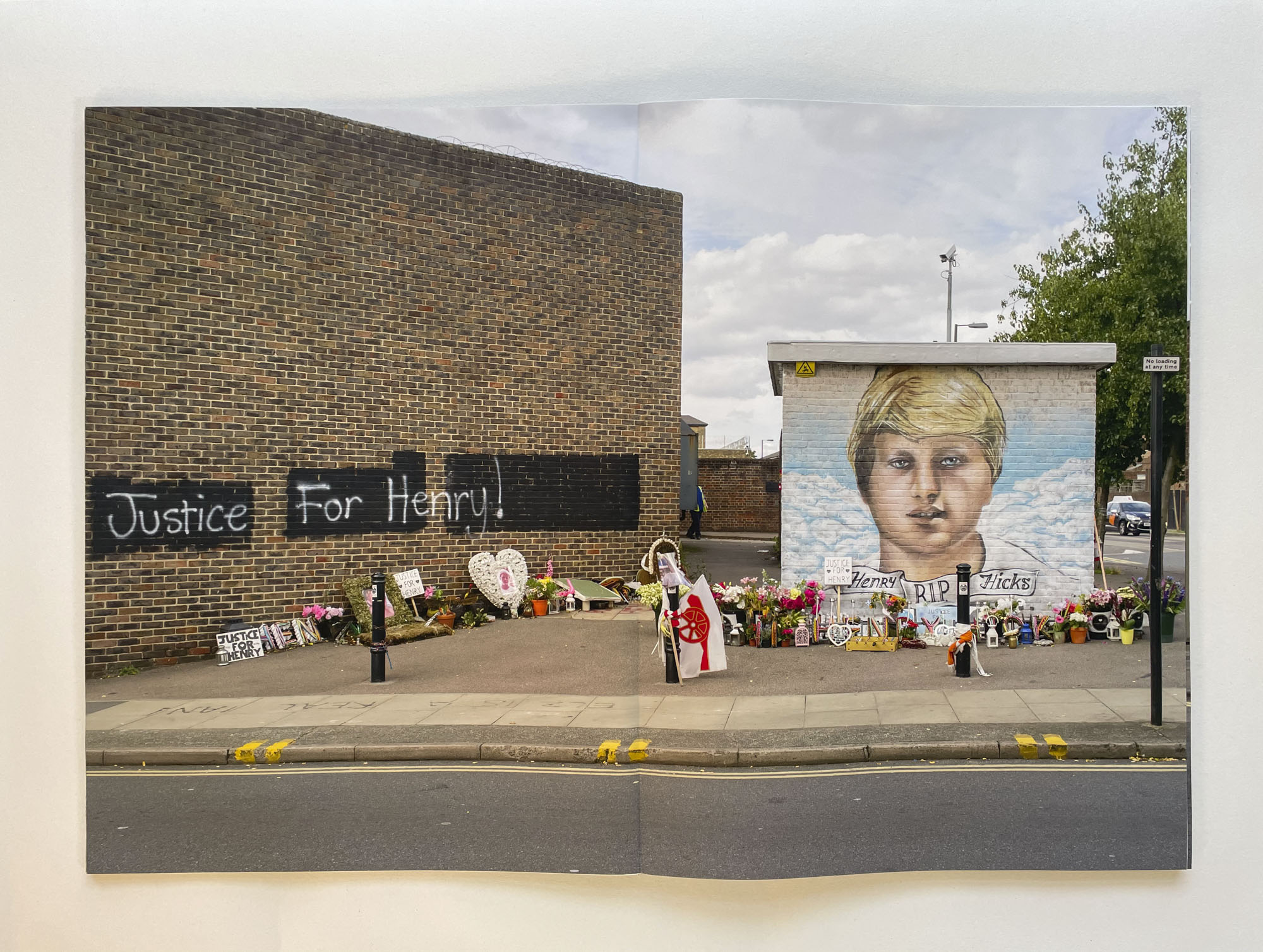 a street memorial with many bouquets of flowers, a painting of the dead boys face against blue sky and on another wall the words Justice for Henry!