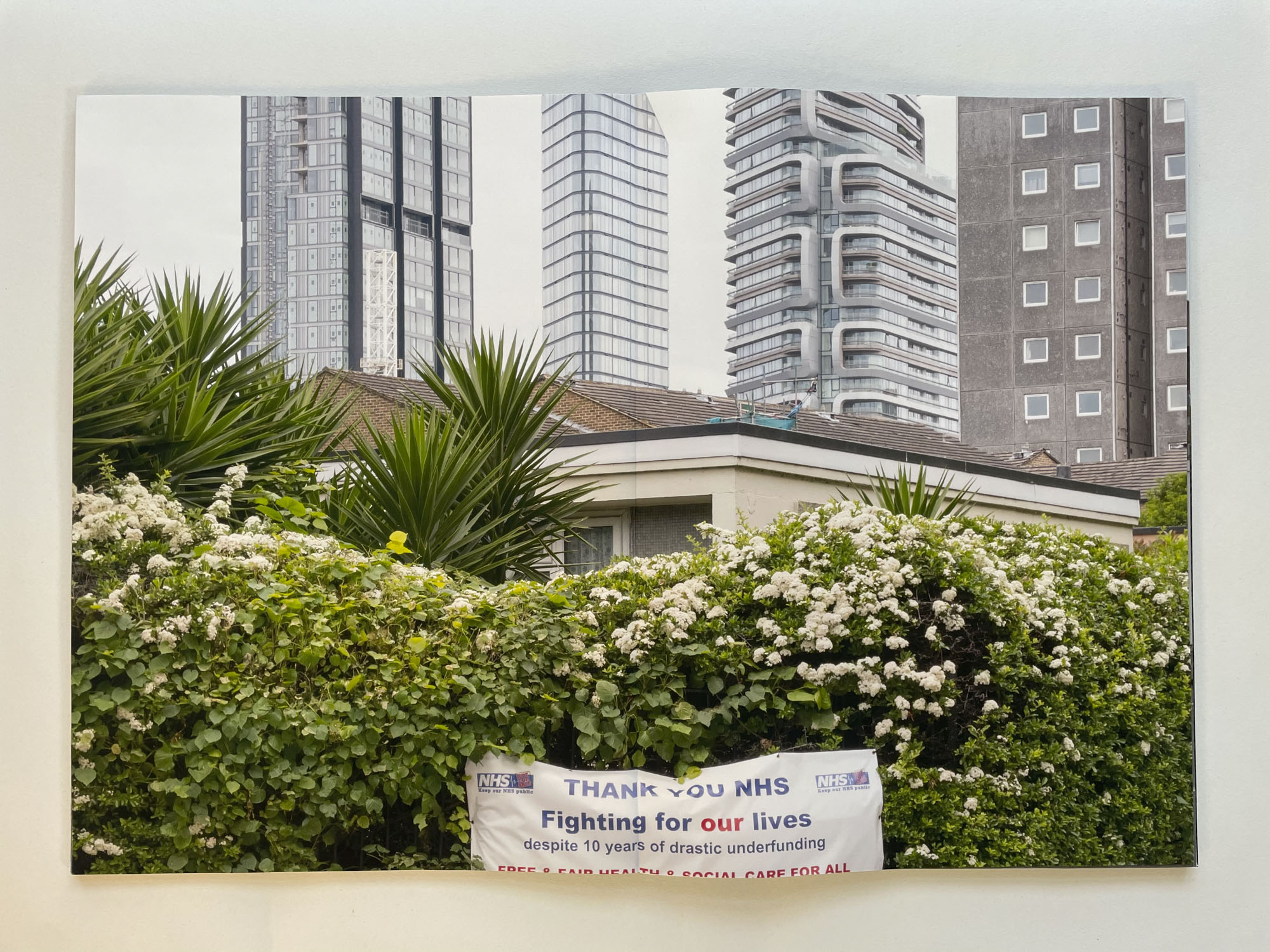 in the foreground against a flowering hedge is a banner with the words Thank you NHS. Fighting for our lives despite 10 years of drastic underfunding; behind rise three glass and steel luxury apartment buildings