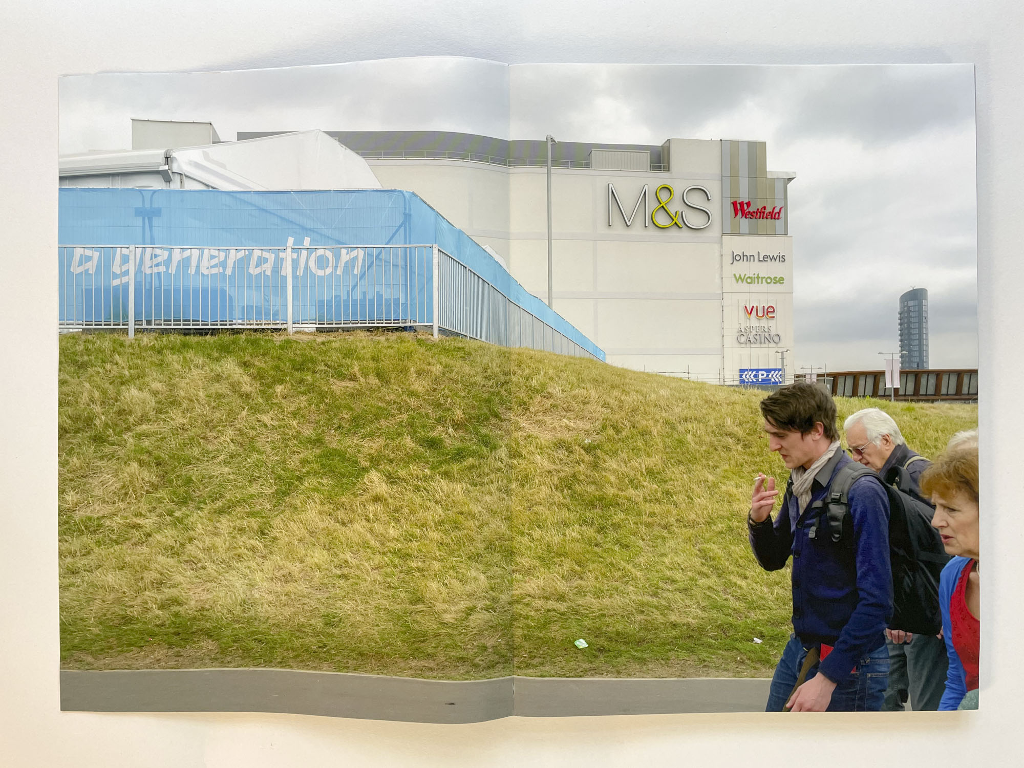 a photograph shows a grass bank with box retail stores in the background, in the right foreground a group of adults walk to the left with worried faces, at the top of the grass bank a hoarding bears the words A generation