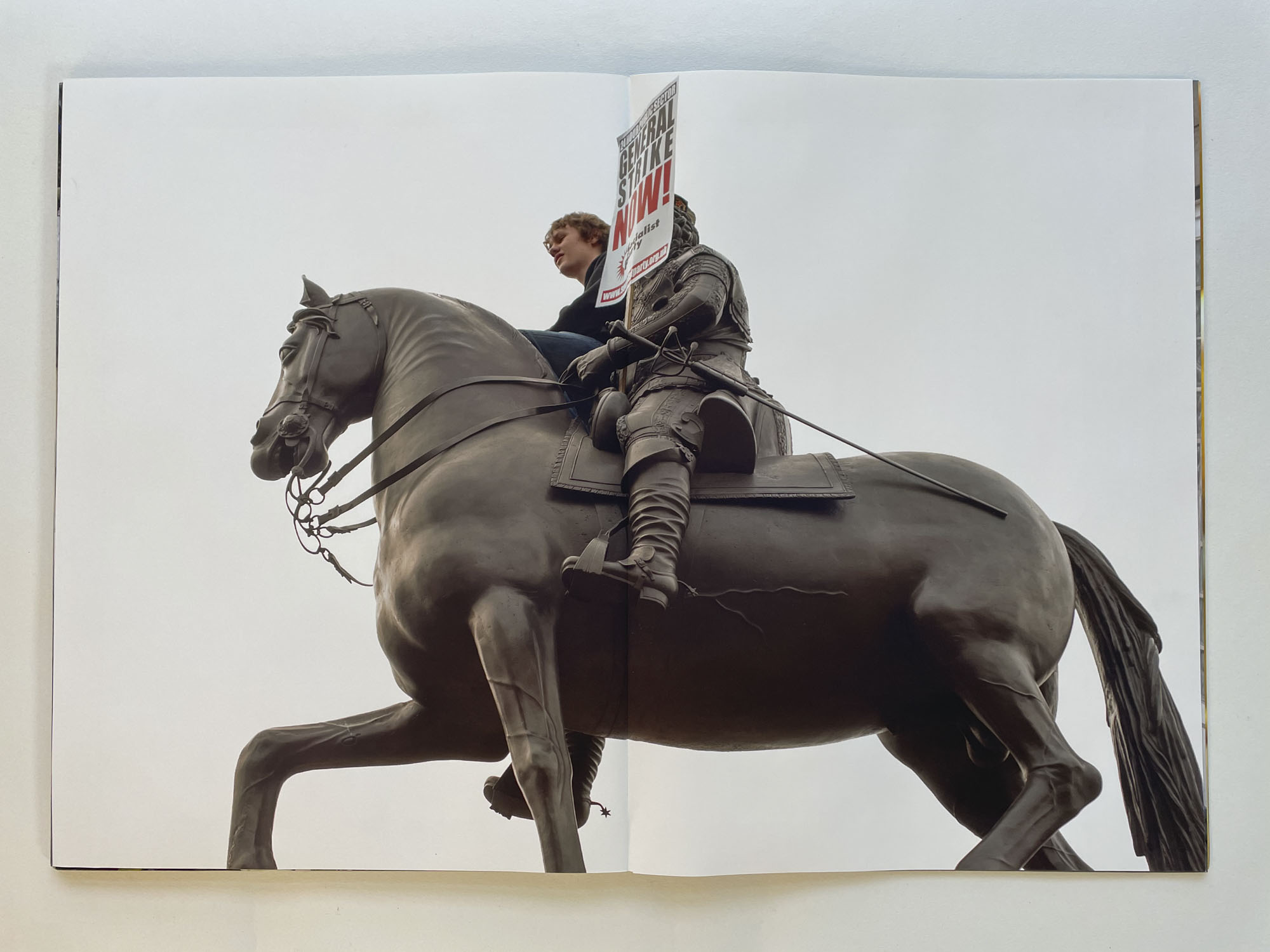 looking up at a statue of a horse and rider, a young man is also sitting on the horse holding a placard that says General Strike Now!