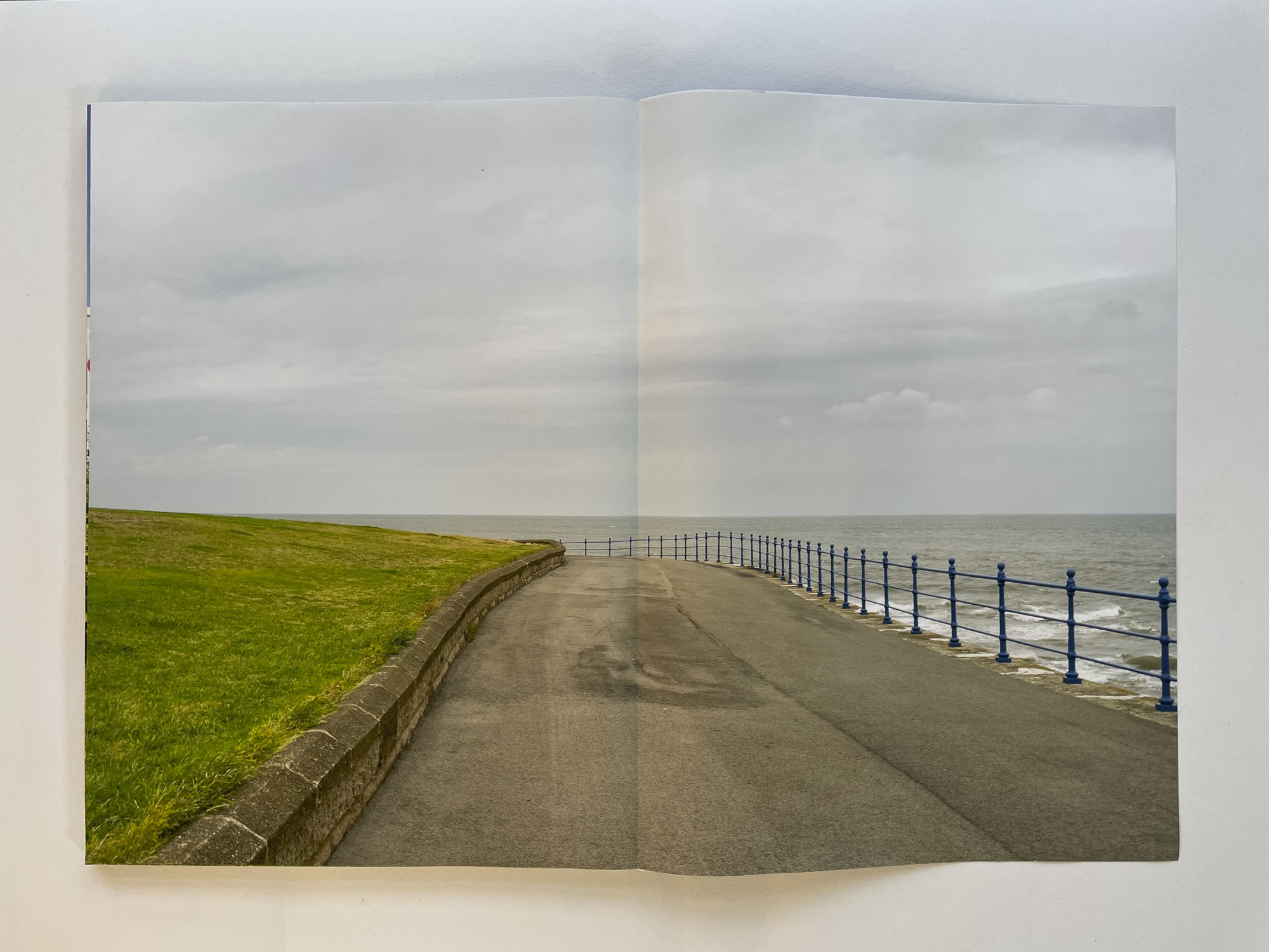 under a grey sky a wide road traces the edge of land, a blue railing separates it from the sea