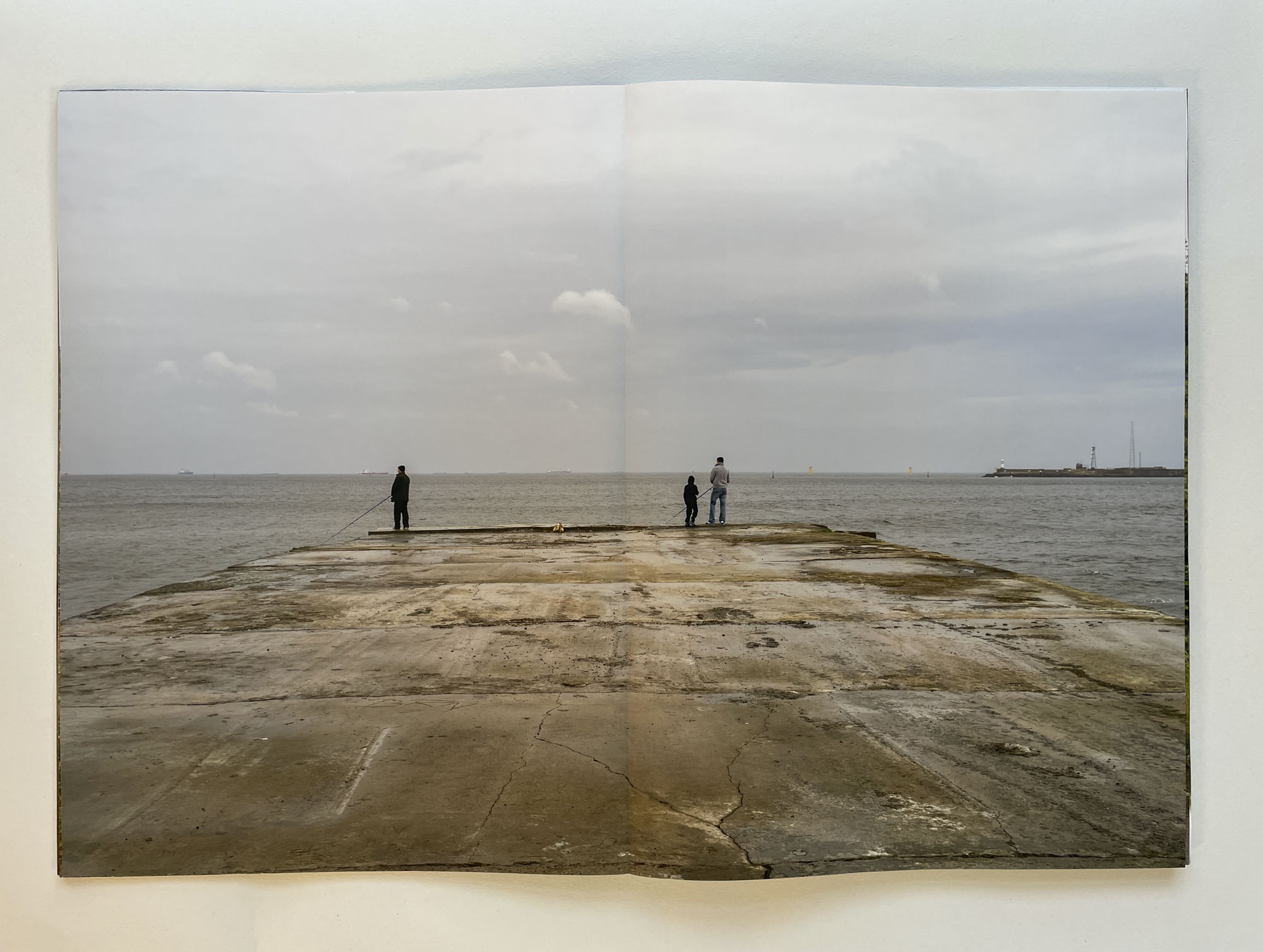 a concrete pier extends into the sea, in the distance are ships, at the end of the pier two men and a boy are fishing