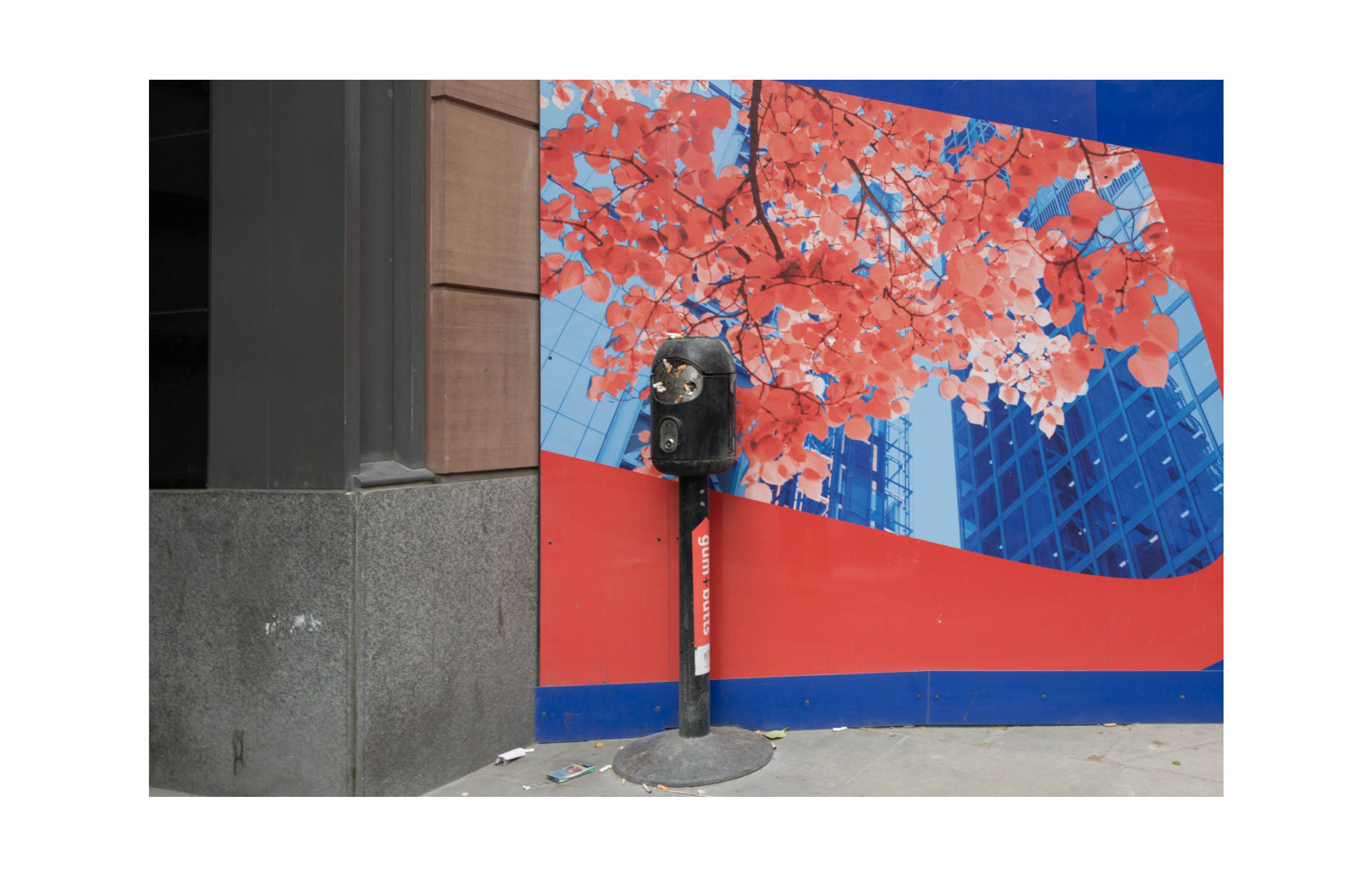 a full to the brim outdoor ashtray stands in front of a bright red and blue hoarding