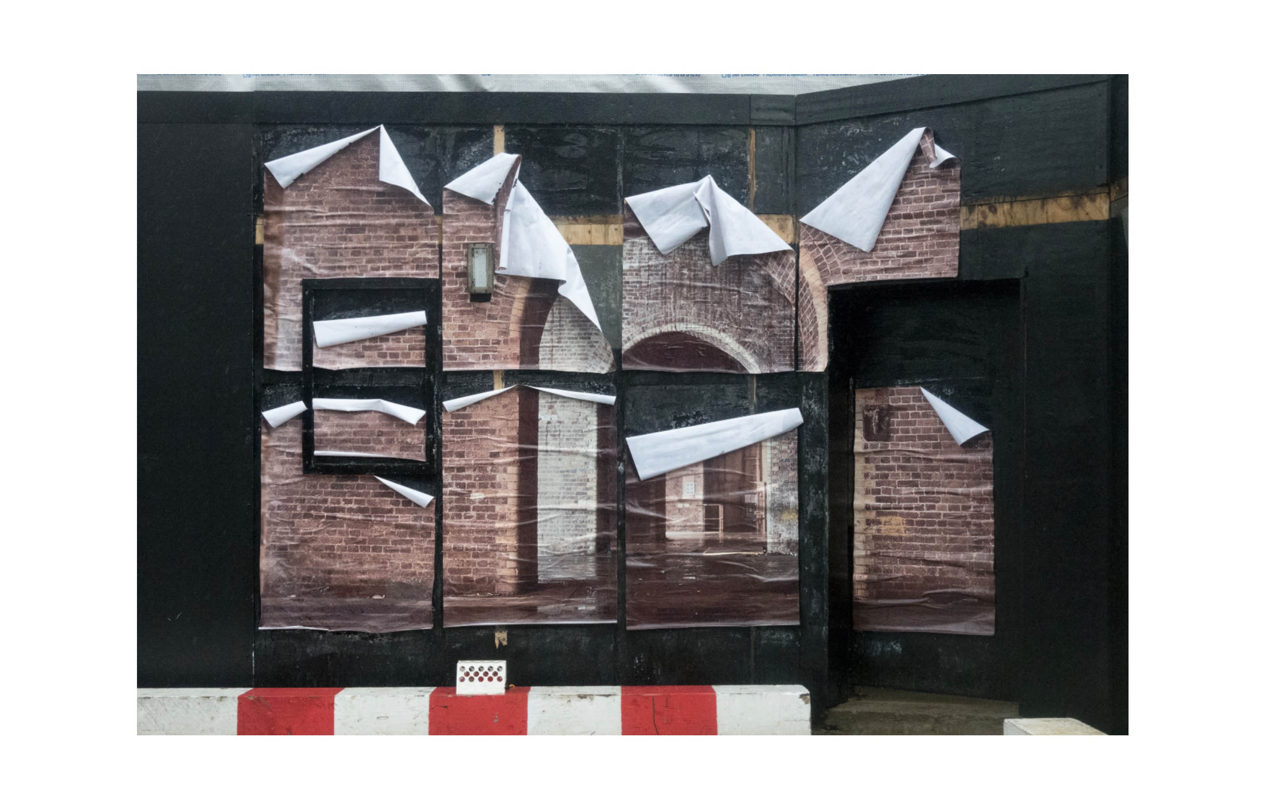 a photograph in eight sections of a brick archway has been pasted onto a black hoarding, rain has caused the pieces to begin to peel away