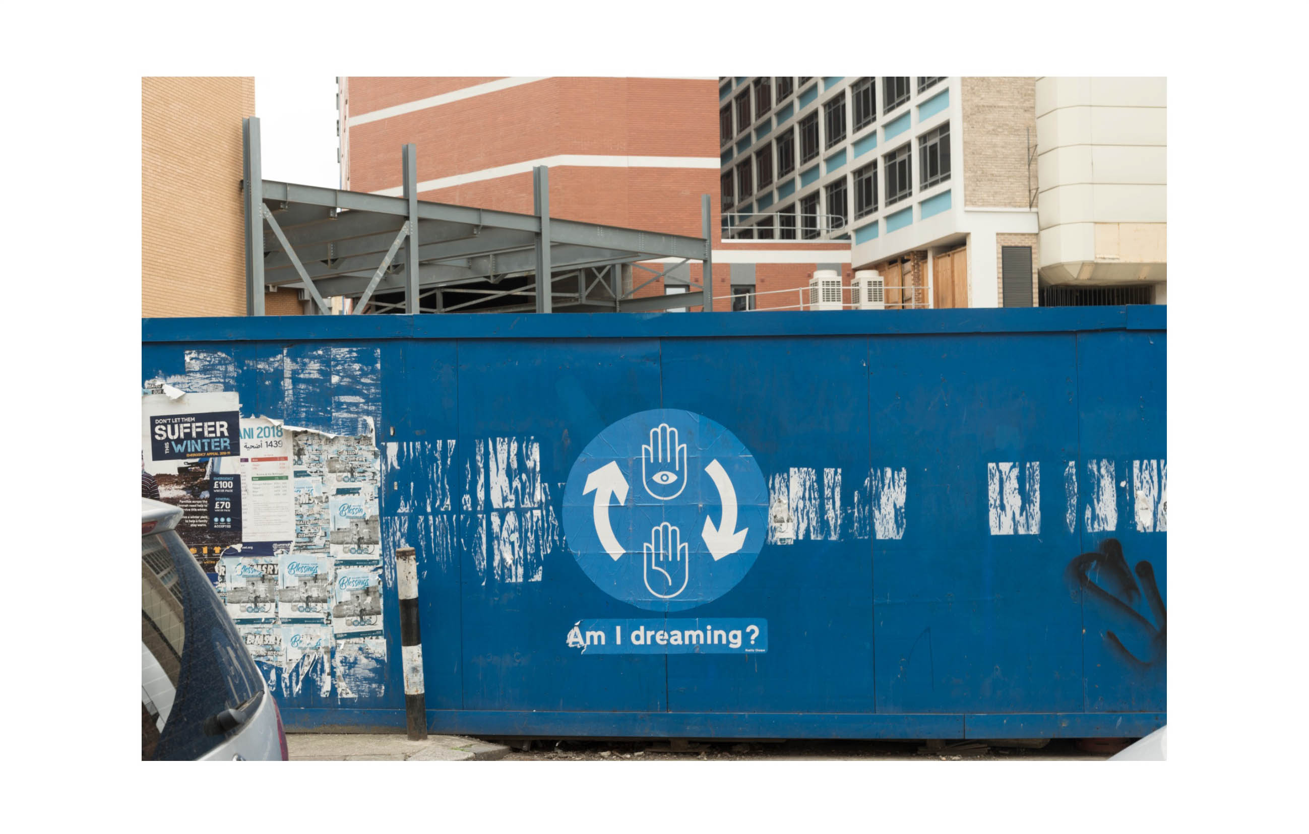 a blue hoarding with a circular poster on which there is a hand with an eye and a hand without one joined by two circular arrows, below this are the words - am I dreaming?