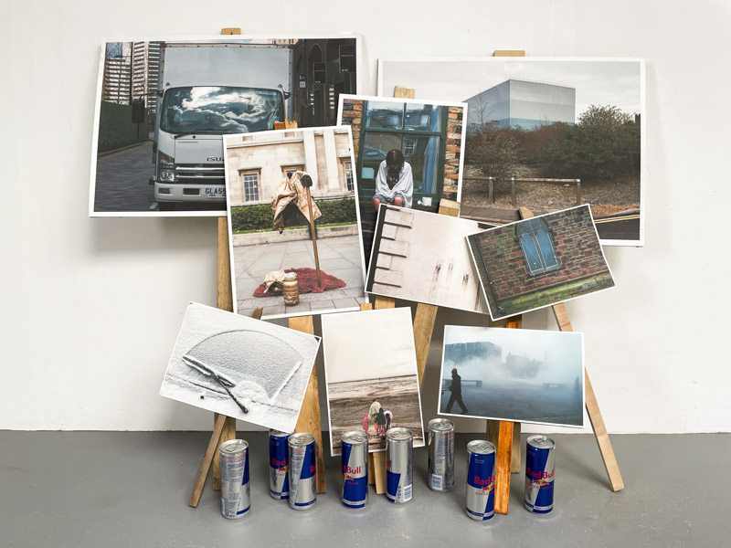 a number of places lean on a wall, the placards bear photographs of disparate subjects, at their feet is a row of unopened Red Bull cans