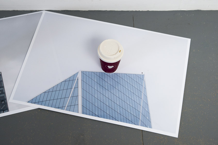 a close-up view of the sixth colour print on glossy paper of the top of a skyscraper against a grey sky, in the centre of which is a Costa coffee cup