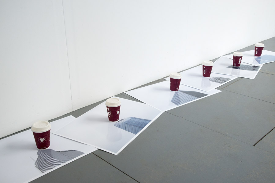 a view of the six large glossy photographic prints of the tops of skyscrapers strewn on the floor below the grid of images, at the centre of each print stands a Costa coffee cup