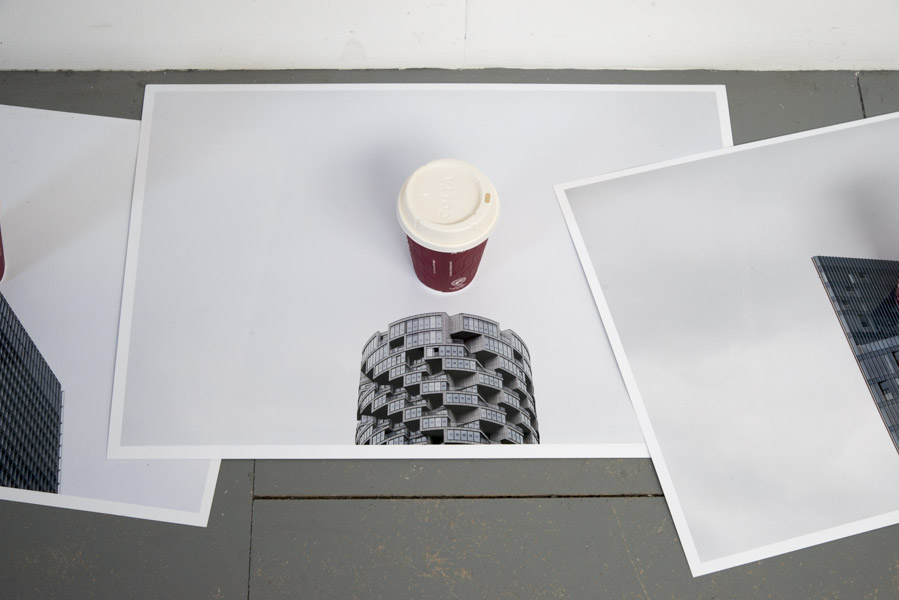 a close-up view of the fourth colour print on glossy paper of the top of a skyscraper against a grey sky, in the centre of which is a Costa coffee cup