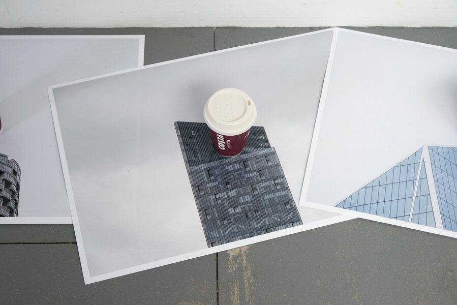 a close-up view of the fifth colour print on glossy paper of the top of a skyscraper against a grey sky, in the centre of which is a Costa coffee cup