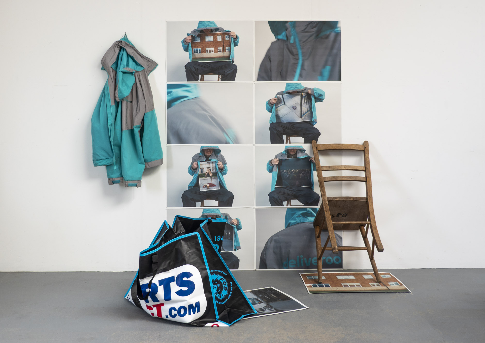 an installation of photographs pinned to a wall in a grid, a Deliveroo jacket, a chair and a Sports Direct bag