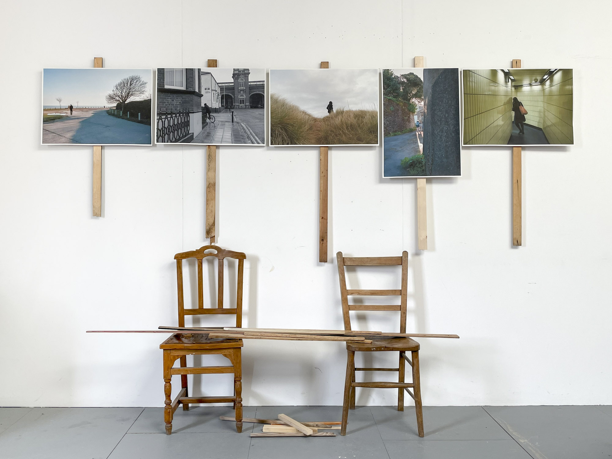 five images of people walking away from the viewer pinned to a wall on poles, two chairs below them have poles balanced between them and on the floor
